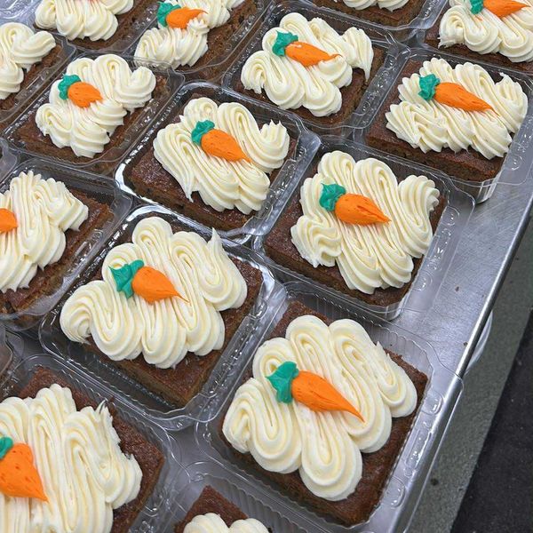Rows of carrot cake squares with piped frosting and little frosting carrots, packed for local pickup
